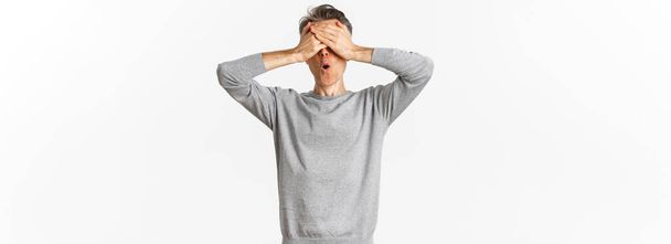 Зображення збудженого хлопця середнього віку в сірому светрі, прикривають очі руками і дивляться здивовані в очікуванні сюрпризу, граючи хованки на білому тлі
. - Фото, зображення