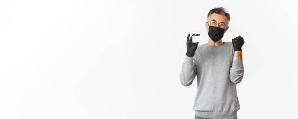 Konzept des covid-19, soziale Distanzierung und Lebensstil. Bild eines attraktiven Mannes mittleren Alters mit medizinischer Maske, Handschuhen und Brille, der Kreditkarte zeigt und jubelt, vor weißem Hintergrund stehend. - Foto, Bild
