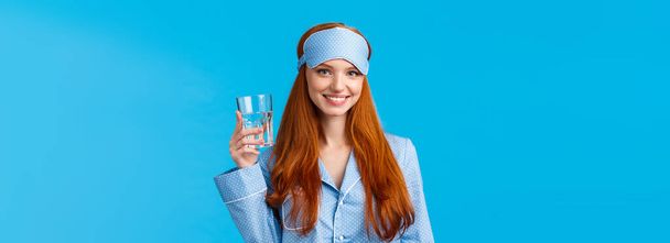 Waist-up shot glamour redhead woman taking care health, eating healthy, drinking water, wearing sleep mask and nightwear, holding glass and smiling energized, standing blue background. - Photo, Image