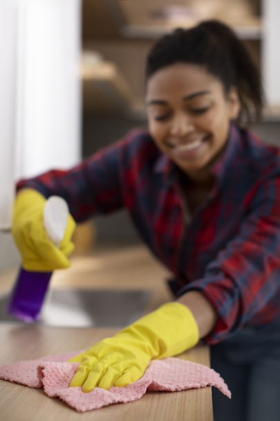 Sonriente millennial africana americana ama de casa en guantes de goma con esponja y spray limpia el polvo de la mesa de madera, disfruta de las tareas domésticas en el interior de la cocina. Perfeccionismo, servicio de limpieza en casa - Foto, Imagen