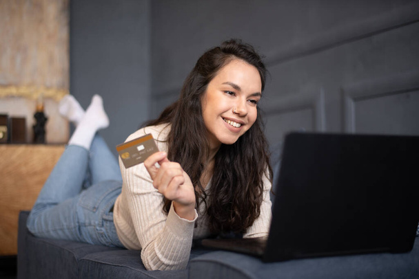 Podekscytowana młoda kobieta robi zakupy online z kartą kredytową i laptopem, leży na kanapie we wnętrzu salonu, wolna przestrzeń. Happy lady surfing web, zakup towarów w sklepie internetowym - Zdjęcie, obraz