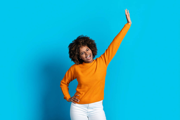 Joyeux heureux belle afro-américaine jeune femme aux cheveux touffus dans une tenue décontractée élégante se déplaçant sur fond bleu studio, levant une main vers le haut et souriant, dame noire dansant seule - Photo, image