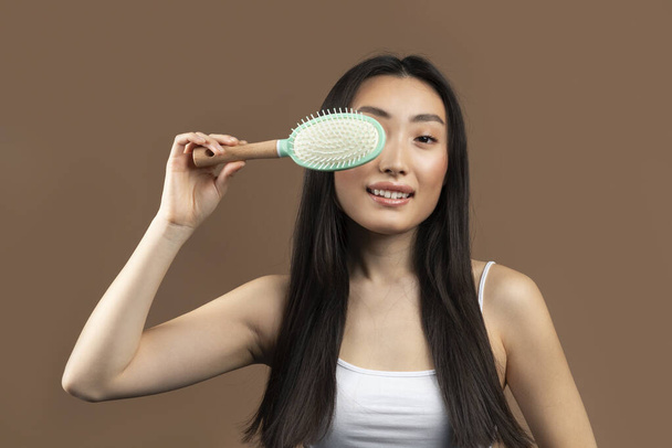 Hiustenhoitokonsepti. Innoissaan korealainen nainen pitkät hiukset peittävät silmän hiusharjalla ja hymyilee kameralle ruskealla taustalla. Hiustyökalujen mainonta - Valokuva, kuva