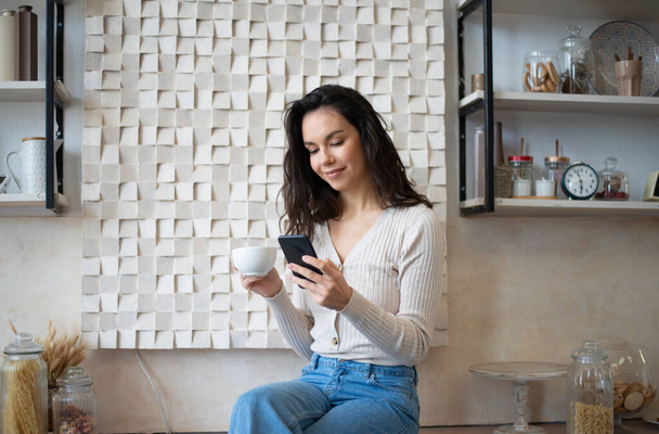 Pozitív hölgy forró reggeli italt iszik és okostelefonon cseveg, üzenetet olvas, a konyha belsejében ül. Közösségi hálózatok, blog és új alkalmazás, hirdetés és jó ajánlat - Fotó, kép