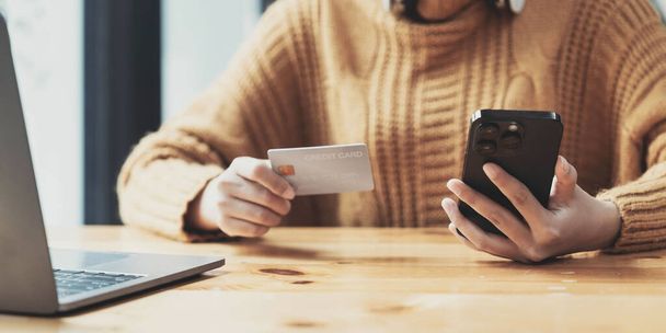 Vonzó ázsiai nő ül az asztalánál, kezében egy okostelefon és egy hitelkártya. Egy közeli kép. Online fizetés, hitelkártyás fizetés, mobil banki szolgáltatások, online vásárlás, készpénz nélküli társadalom... - Fotó, kép
