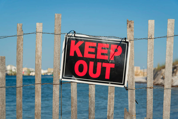 Rot-schwarzes Keep Out-Schild an einem Zaun aus Draht und Holzlatten in Destin, Florida. Großaufnahme eines Schildes an einem Zaun vor blauem Wasser und Himmel. - Foto, Bild