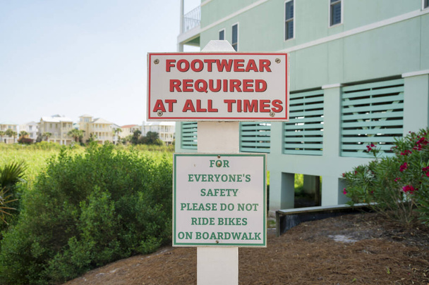 Firme con el calzado requerido en todo momento y por favor no conduzca bicicletas en el paseo marítimo en Destin, FL. Señalización fuera del edificio verde claro cerca de las hierbas altas en el fondo. - Foto, imagen