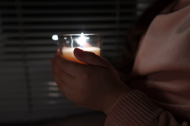 A girl in a dark room near the window holds a candle in a glass glass in her hands (close-up). Power outage concept. Blackout. Energy crisis. Destruction of infrastructure - Photo, Image