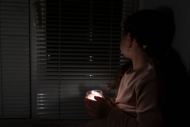 A girl in a dark room near the window holds a candle in a glass in her hands. Power outage concept. Blackout. Energy crisis. Destruction of infrastructure - Photo, Image