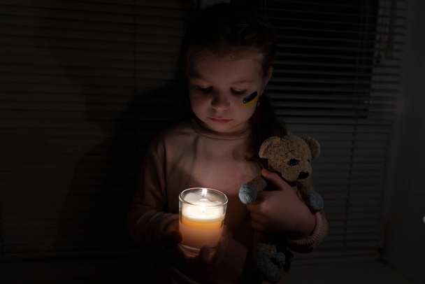 A girl with a painted flag of Ukraine on her cheek in a dark room holds a candle and a teddy bear in her hands. Power outage concept. Blackout. Energy crisis. Destruction of infrastructure - Photo, Image