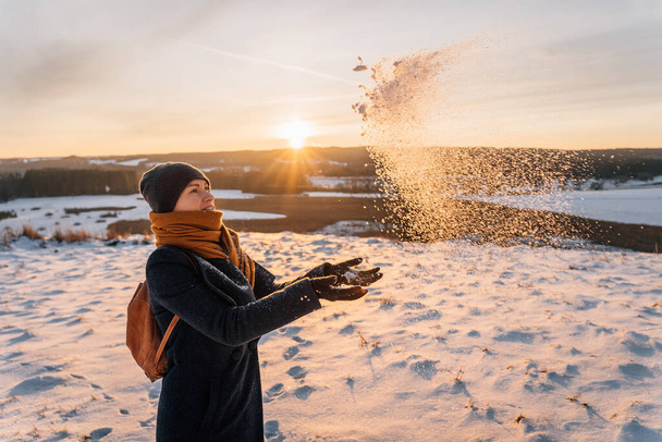 暖かい服を着た冬の女性が夕日を背景に手のひらと雪を投げかける. - 写真・画像