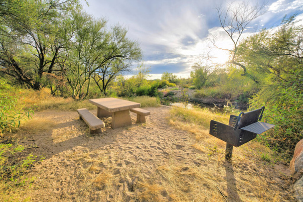 Sabino Canyon State Park, Tucson, Arizona- área de acampamento com churrasqueira de madeira e vista de um riacho. Há uma grelha no chão à direita e mesa de concreto com assentos contra o fundo do céu brilhante. - Foto, Imagem