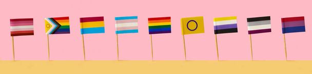 algunas banderas LGBTIQA diferentes unidas a postes parados uno al lado del otro sobre una superficie amarilla, sobre un fondo rosa, en un formato panorámico para usar como banner web - Foto, Imagen