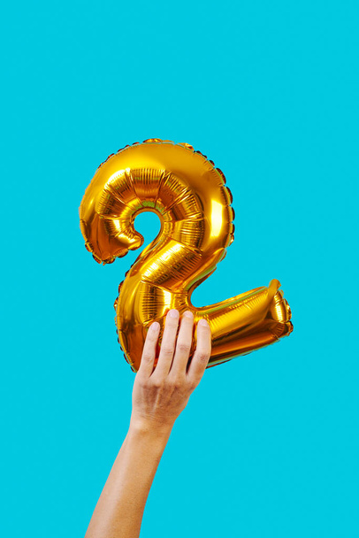 άνθρωπος κρατά ένα χρυσό μπαλόνι στο σχήμα του αριθμού 2 σε ένα μπλε φόντο - Φωτογραφία, εικόνα