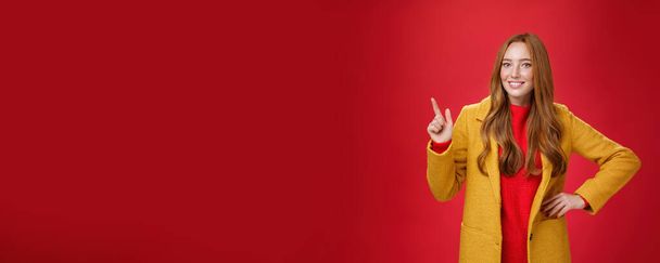 Encantadora joven de abrigo amarillo agarrada de la mano en la cintura y apuntando a la esquina superior izquierda, sonriendo ampliamente como mostrando o promoviendo una propuesta interesante sobre el fondo rojo, de buen humor. - Foto, Imagen