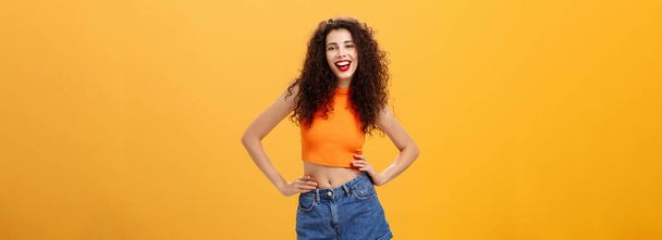 Stylish confident and awesome woman. ready lit party standing in trendy cropped top and denim shorts with hands on waist sticking out tongue and smiling joyfully posing over orange background. - Photo, Image