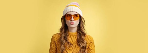 Stílusos fiatal glamour figyelmes lány, hogy a kiszáradás összecsukható ajkak buta gondolkodás termék kiválasztása elfordul visel napszemüveg kalap meleg pulóver élvezi nyaralás sípálya, sárga háttér. - Fotó, kép