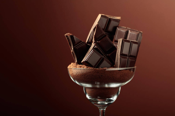 Broken black chocolate bar in glass with cocoa powder. Chocolate on a brown background with copy space. - Photo, Image