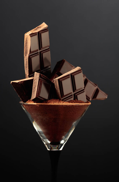 Zerbrochene schwarze Schokoladentafel im Glas mit Kakaopulver. Schokolade auf schwarzem Hintergrund. - Foto, Bild