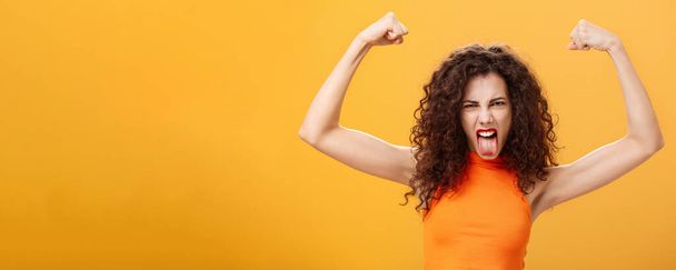 Cintura di femmina caucasica fredda e audace in top arancione con tatuaggio sul braccio accigliato che rende divertente il viso che sporge la lingua alzando le mani mostrando muscoli che provano potenza e forza. Copia spazio - Foto, immagini