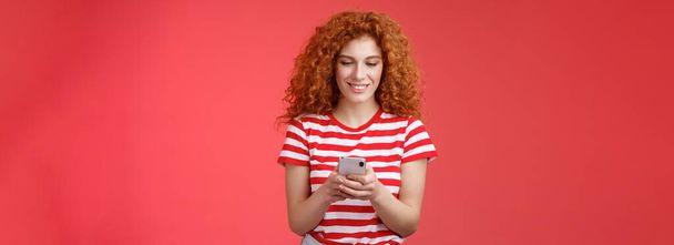 Έλεγχος χρόνου στα μέσα κοινωνικής δικτύωσης. Όμορφα χαλαρή ευτυχισμένη μοντέρνα κοκκινομάλλα γυναίκα σγουρά χτένισμα κρατήστε το smartphone ματιά οθόνη του τηλεφώνου διασκεδάζει χαμογελώντας χαρούμενος γραπτών μηνυμάτων φίλο ρίξει κόμμα το συντομότερο. - Φωτογραφία, εικόνα