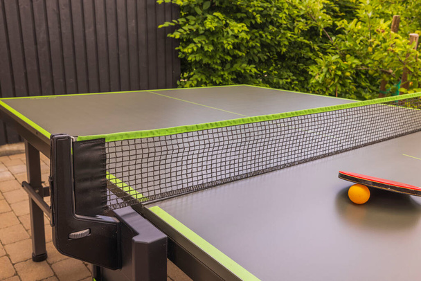 Pohled na pingpongovou raketu a oranžový plastový míček na zeleném tenisovém stole ve dvoře vily. Koncept aktivního životního stylu. Švédsko. - Fotografie, Obrázek