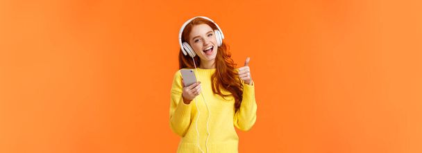 Приваблива руда дівчина отримує різдвяний подарунок нові круті навушники, слухаючи музику в навушниках і показуючи великий палець, тримаючи смартфон, насолоджуючись музикою, співаючим і посміхаючись, помаранчевим фоном
. - Фото, зображення