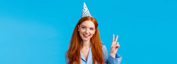 Gelukkige en enthousiaste vrouwelijke mooie roodharige vrouw met lang krullend haar in nachtkleding, het dragen van verjaardagspet tonen vrede teken en vrolijk glimlachen, vieren b-dag, blauwe achtergrond. - Foto, afbeelding