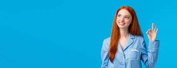 夢のような創造的なかわいい赤毛の女性は、ナイトウェアを着て、新しい衛生製品を承認し、青い背景に立って、良いジェスチャーを示して、幸せと安心を感じています. - 写真・画像