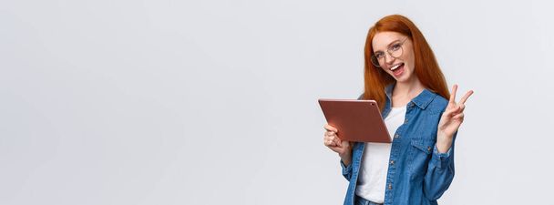 Sassy and cute, carefree happy redhead woman in glasses, denim shirt, showing peace sign and smiling camera cheerful as holding digital tablet, watching vlog online, working from distance. - Photo, Image