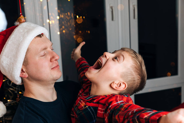 Vater und Sohn in roten Pyjamas neben dem Weihnachtsbaum und umarmen sich. Frontansicht - Foto, Bild