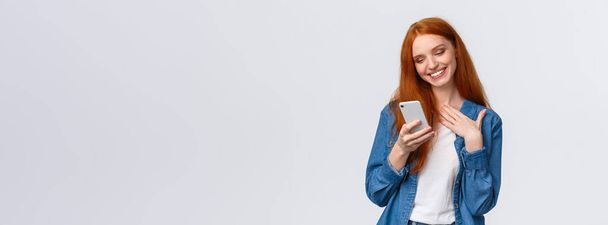 Oh so cute. Touched and pleased alluring redhead girlfriend receive love emoji, lovely message, texting, chatting boyfriend, holding smartphone, touch chest and smiling mobile display. - Photo, Image