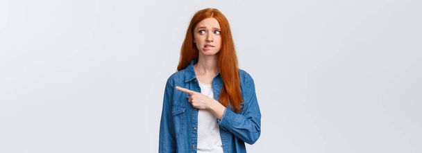 Awkward and nervous cute timid redhead teenage girl having fear or embarrassing thing say, pointing looking right anxious, biting lip and frowning, confessing in crime, feel guilty. - Photo, Image