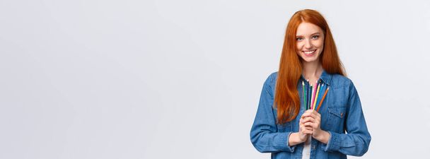 Time to brighten life. Attractive confident charismatic redhead girl in denim shirt, looking camera with pleased smile, holding colored pencils, standing white background drawing, attend courses. - Photo, Image