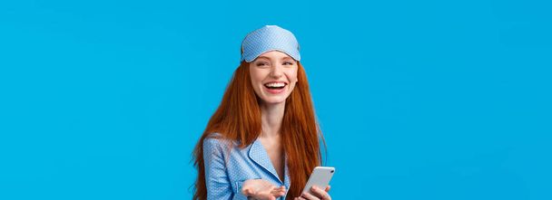 Glückliche und charismatische rothaarige Frau in Nachthemd, Schlafanzug und Schlafmaske, lachend und sagend, was für ein süßes Lächeln, Smartphone sprechende Mitbewohnerin, stehend blauer Hintergrund. - Foto, Bild