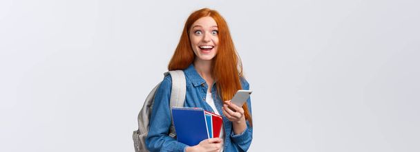 Opgewonden en gelukkig, opgelucht schattig roodharige vrouwelijke student met rugzak, notities in notebook en smartphone, op zoek vrolijke camera, sms 'en, berichten vrienden, witte achtergrond. - Foto, afbeelding
