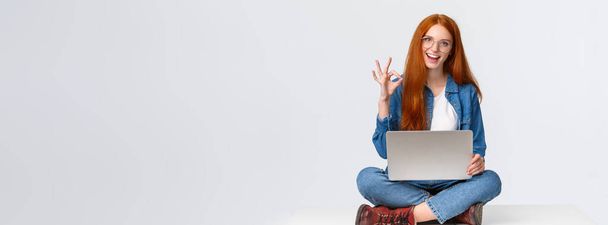 Dont worry, all done, everything under control. Confident happy and relaxed redhead girl saying all okay, show ok gesture while sit on floor with legs crossed, holding laptop, white background. - Photo, Image