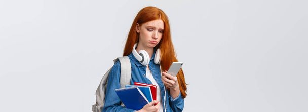 Silly sad and gloomy pouting girl with red foxy hair, frowning sighing and looking with regret smartphone screen, reading troublesome message, feeling bad, stand with backpack and notebooks. - Photo, Image