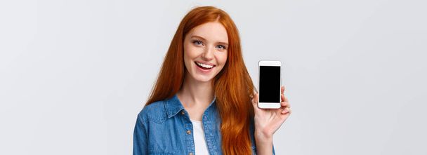 Close-up retrato moderno millennial linda pelirroja con ojos azules, consejos obtener nueva aplicación fresca, mostrando la pantalla del teléfono inteligente y sonriendo, juego de publicidad o aplicación de filtro de fotos
. - Foto, Imagen