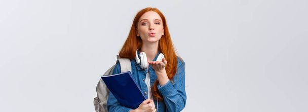 Sevimli ve hassas, şehvetli bir üniversiteli kız, kızıl saçlı şımarık bir öğrenci, hava öpücüğü gönder, kameraya tutkuyla üfle, sırt çantası ve klasörü tut.. - Fotoğraf, Görsel