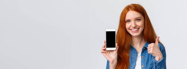 Közelkép portré elégedett csinos vörös hajú nő segítségével alkalmazás, töltse le, reklám fotó app, online áruház, show smartphone kijelző és hüvelykujj-up, mint. - Fotó, kép