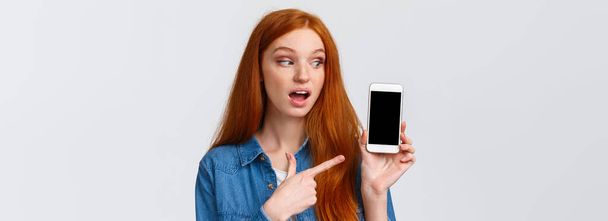 Curioso, si chiedeva abbastanza rossa femmina caucasica che mostra le foto da vacanza, indicando display smartphone sbirciando sullo schermo, in piedi divertito su sfondo bianco, raccomandare app. - Foto, immagini