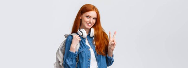 Прекрасна кокетка і нахабна руда дівчина коледжу, носити навушники над шиєю і рюкзаком, показуючи знак миру і усміхнену усміхнену камеру, що стоїть щасливо на білому тлі насолоджуйтесь студентським життям
. - Фото, зображення