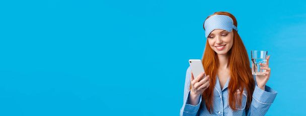 Glückliche und entspannte attraktive rothaarige Frau, die Smartphone und Glaswasser in der Hand hält, freudig und erfreut lächelt, als sie schöne Nachrichten aus der mobilen Anwendung liest, blauer Hintergrund. - Foto, Bild