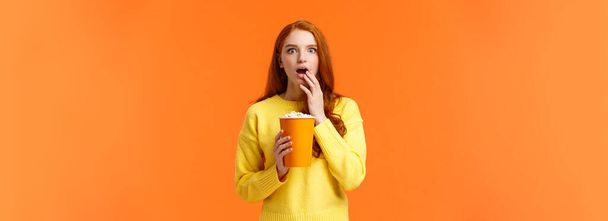 schockiert und interessiert, unterhaltsames rothaariges Mädchen, das einen tollen neuen Film auf der Großleinwand anschaut, Popcorn isst, erstaunt den Mund aufreißt, von cooler Filmhandlung erschrocken ist, orangefarbener Hintergrund. - Foto, Bild