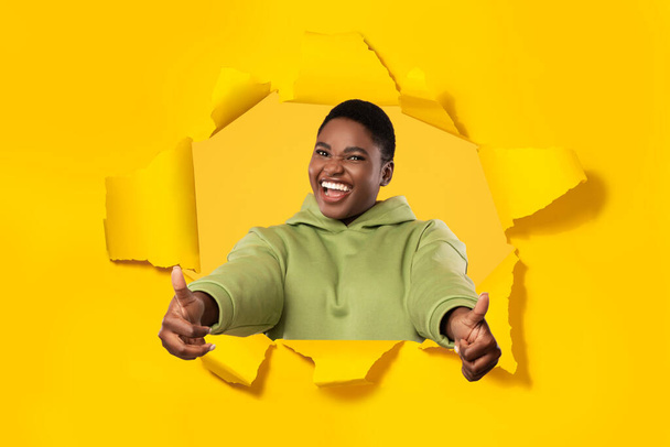 Excited black plus size woman gesturing thumbs up, stretching arms to camera and smiling through hole in torn yellow paper. Lady approving great offer. I like it concept - Photo, Image