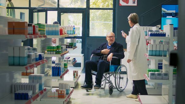 Ειδικός υγείας που μετρά τη θερμοκρασία στον άνθρωπο με αναπηρία, χρησιμοποιώντας θερμόμετρο με άτομο σε αναπηρική καρέκλα. Άνδρας πελάτης που αγοράζει συνταγογραφούμενα φάρμακα σε φαρμακείο φιλικό προς την αναπηρία. - Φωτογραφία, εικόνα