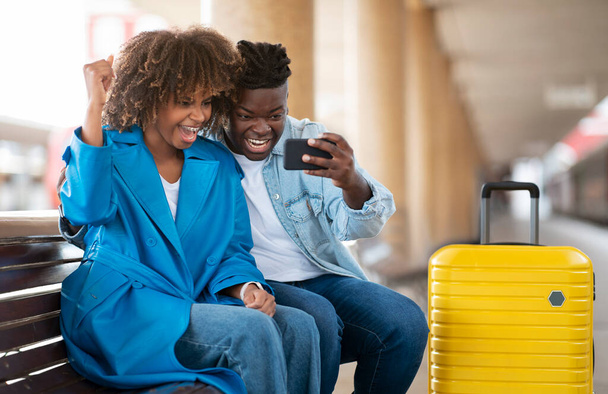 Pareja negra llena de alegría celebrando el éxito con el teléfono inteligente mientras se sienta en el banco en la estación de tren, cónyuges afroamericanos felices ganando juegos en línea juntos mientras esperan tren, espacio libre - Foto, Imagen