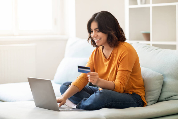 Mosolygó fiatal arab nő, hogy az online fizetés Laptop és a hitelkártya, miközben ül a kanapén otthon, Boldog Millenniumi közel-keleti nő élvezi a vásárlás az internetről, másolás tér - Fotó, kép