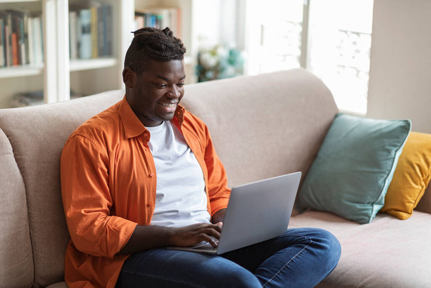 Δροσερό χαμογελαστό παχουλό χιλιετή μαύρο άντρα με πλεξούδες σε casual στολή κάθεται στον καναπέ στο άνετο σαλόνι με τον υπολογιστή στην αγκαλιά του, διευθυντής περιεχομένου που εργάζονται από το σπίτι, αντιγραφή χώρου - Φωτογραφία, εικόνα